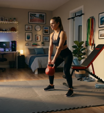 Mulher treinando com kettlebell em um quarto adaptado como academia em casa, com piso de EVA, banco regulável e halteres ao fundo.